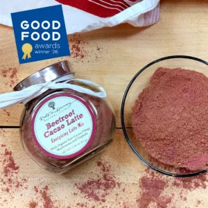 Roots in Harmony Beetroot Cacao Latte in a small glass jar. It is sitting on a light brown table with some in a glass next to it and a little powder sprinkled on the table all around it.