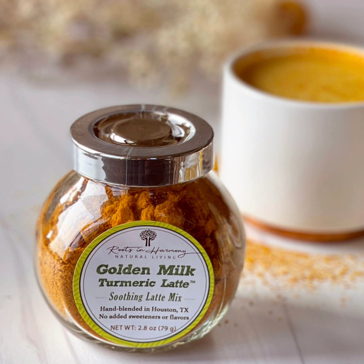 Roots in Harmony brand Golden Milk Turmeric Latte Organic Soothing Mix in a glass jar. It is sitting on a table in front of a white mug filled with a deep turmeric-yellow colored latte.